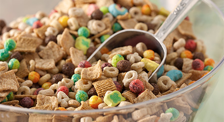 Assorted dry cereal in a large clear bowl with metal scoop.