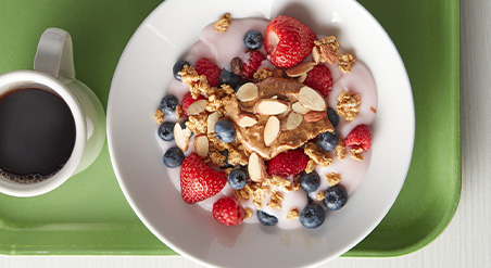  White bowl containing yogurt, granola, and assorted berries. On a bright green cafeteria tray with white mug of coffee.