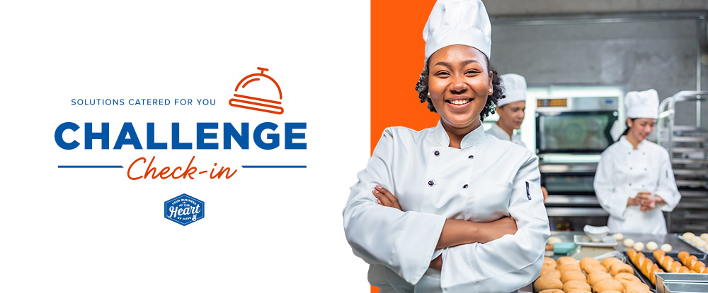 Challenge check-in text in blue and orange on a white background. Icon of a concierge bell in orange. Black female chef in white chef’s coat and hat smiles and stands with her arms crossed. Commercial kitchen scenery in background with one other person working.