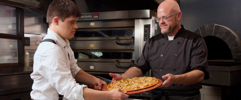 Two men holding a paddle with pizza over a countertop