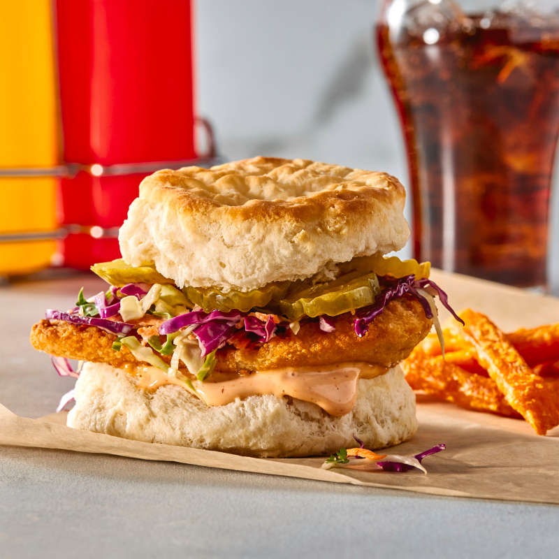A crispy fried chicken sandwich on a fluffy biscuit, topped with coleslaw, pickles, and a creamy orange sauce. It is served on parchment paper with a side of fries and a drink in the background. ortcakes with whipped cream and strawberries, topped with a whole strawberry, against a blue background 