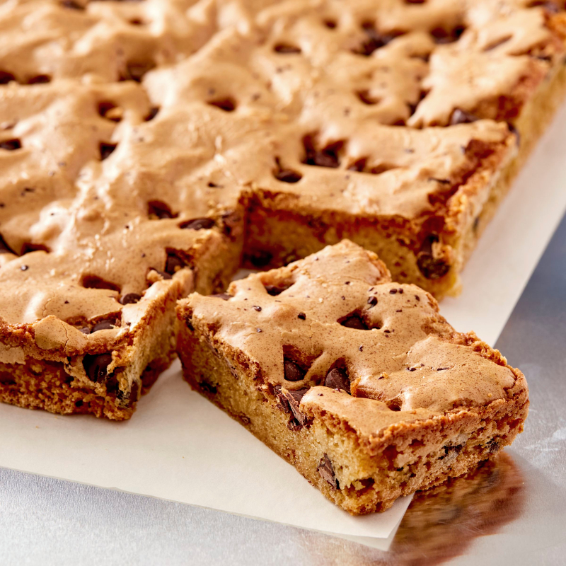 A large sheet of golden brown chocolate chip blondies with a crinkled top, with one piece cut out and resting on parchment paper in the foreground.