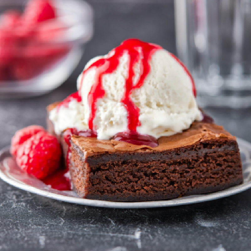 Two fudgy chocolate brownies with crinkly tops and visible chocolate chips are stacked on a metal spatula, resting on a rustic wooden table with scattered chocolate pieces.